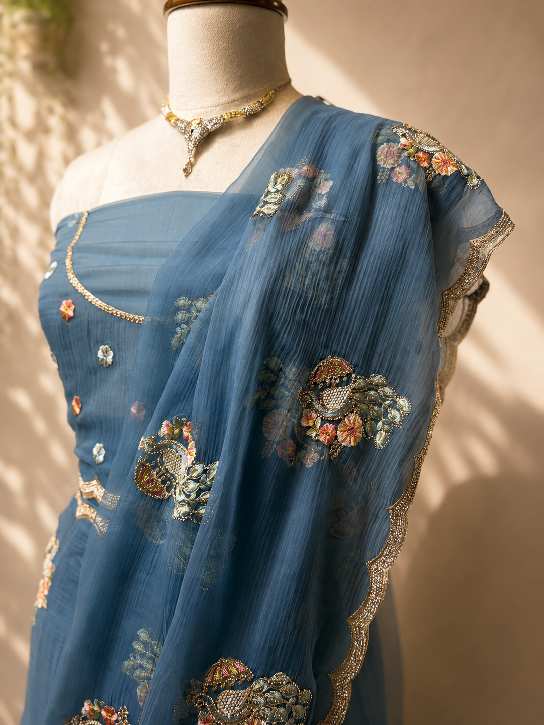 Dusty blue chiffon saree with delicate floral embroidery and scalloped border draped elegantly on a mannequin against a soft beige studio background.
